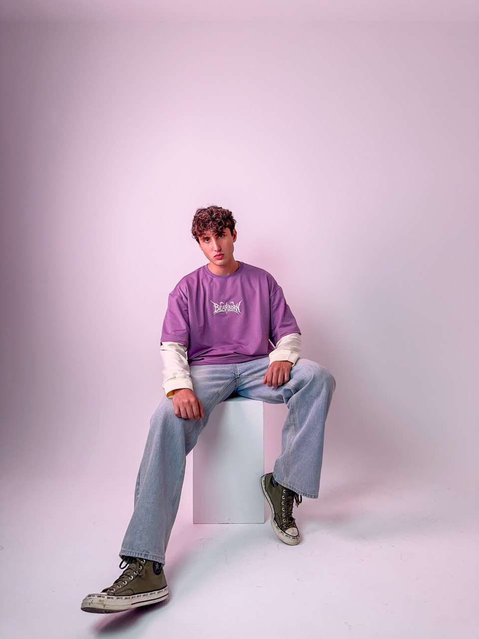 A model seated on a studio block wearing the purple Y2K Double Layer Tee with bone sleeves