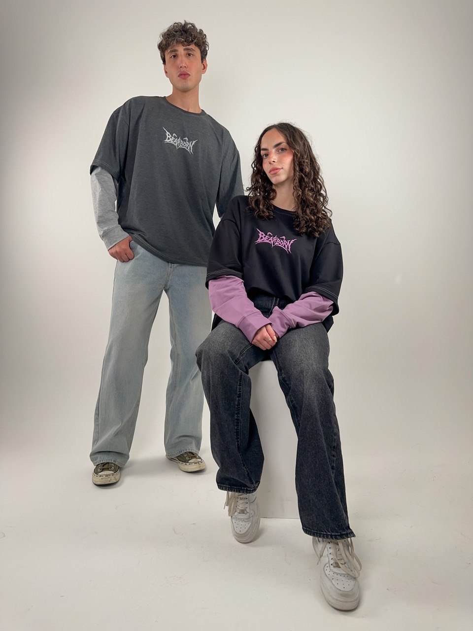 Two models in the Y2K Double Layer Tee — charcoal with grey sleeves and black with lavender sleeves — in a white studio